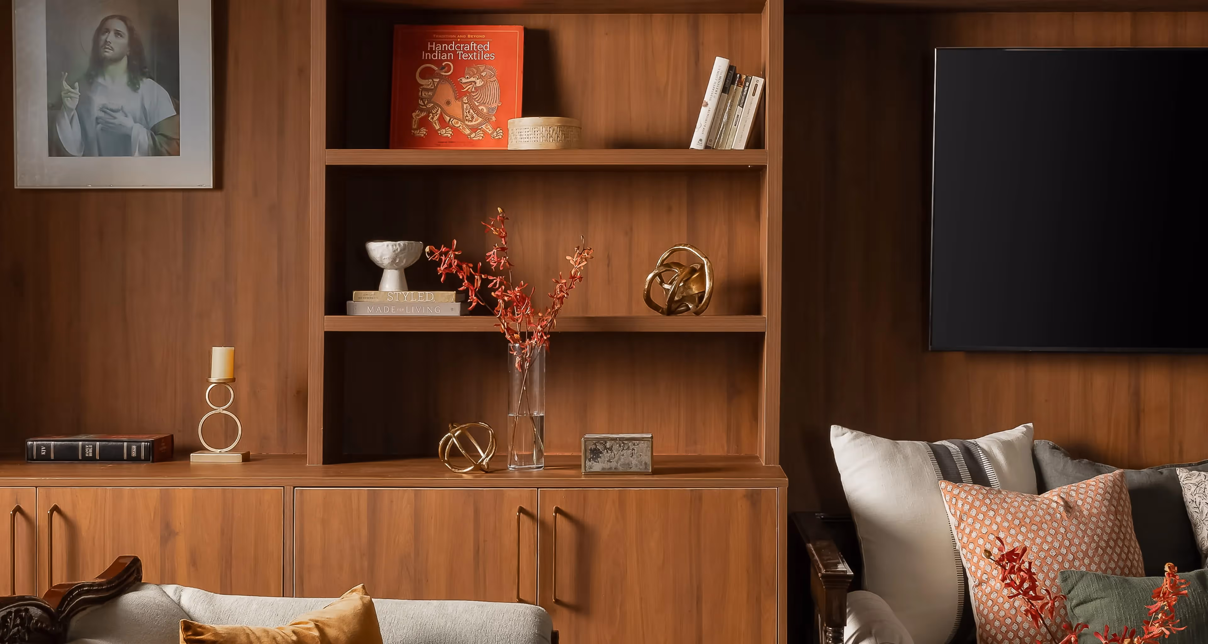 Cozy living room with wooden shelves holding books, decorative items, and a vase with red flowers, adjacent to a TV and cushioned seating.