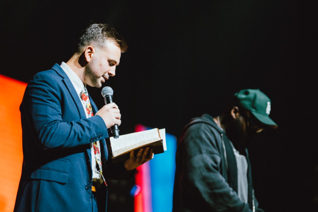 A man in a suit reads from an open Bible while speaking into a microphone onstage, with another man standing beside him in a hoodie and cap against a dark, colorful backdrop.