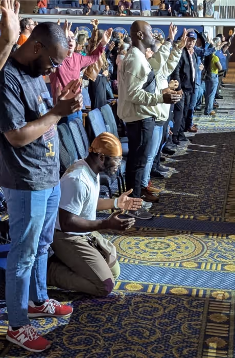 A large group of attendees stands with raised hands in worship while one man kneels at the front in prayer, illuminated by stage lighting in a packed auditorium.