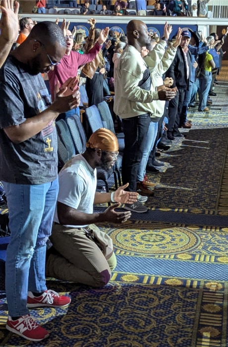 A large group of attendees stands with raised hands in worship while one man kneels at the front in prayer, illuminated by stage lighting in a packed auditorium.