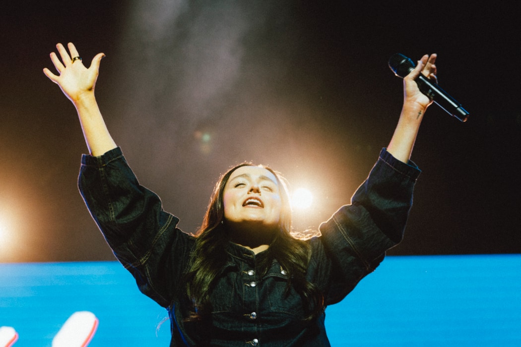 A worship artist Kate stands onstage with arms raised and a microphone in hand, framed by bright backlighting that emphasizes the emotion and energy of the moment.