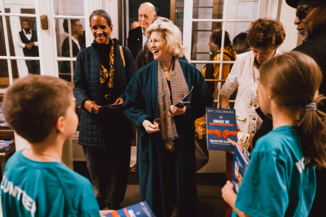 A group of attendees enters an event space smiling as young volunteers in teal shirts hand out “Awake DC” journals; the atmosphere feels warm, welcoming, and energetic.