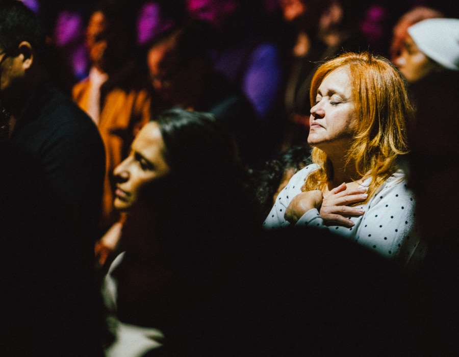 A woman stands in a dimly lit crowd with her eyes closed and hands crossed over her chest, surrounded by others in quiet worship as colored stage lights cast a warm glow.