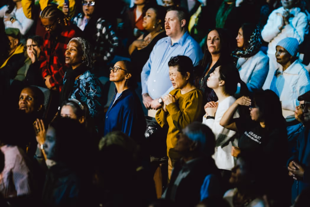 A diverse crowd stands together in a packed auditorium, many with eyes closed, hands raised, or clasped in prayer, creating a unified atmosphere of worship and reflection.