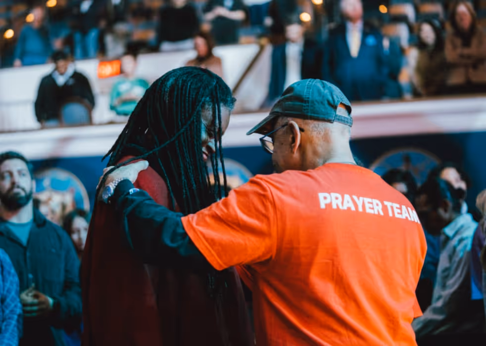 A member of the prayer team in a bright red shirt places a hand on someone’s shoulder, praying with them amid a crowded auditorium filled with people worshipping and watching.