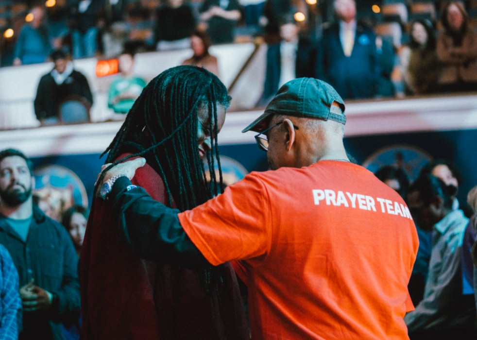 A member of the prayer team in a bright red shirt places a hand on someone’s shoulder, praying with them amid a crowded auditorium filled with people worshipping and watching.
