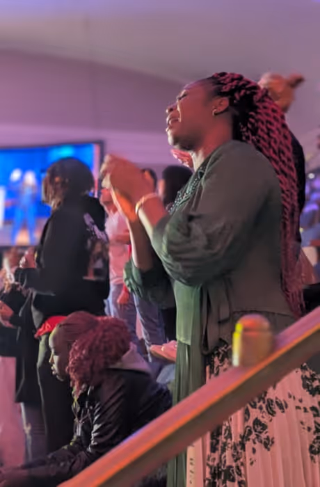 A woman stands near the railing in a dimly lit auditorium, eyes closed and hands lifted in worship, surrounded by others engaged in the moment as colorful stage lighting fills the space.