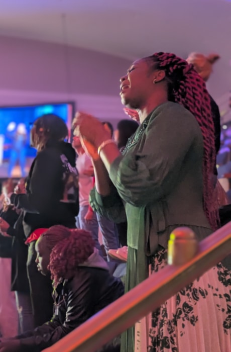 A woman stands near the railing in a dimly lit auditorium, eyes closed and hands lifted in worship, surrounded by others engaged in the moment as colorful stage lighting fills the space.