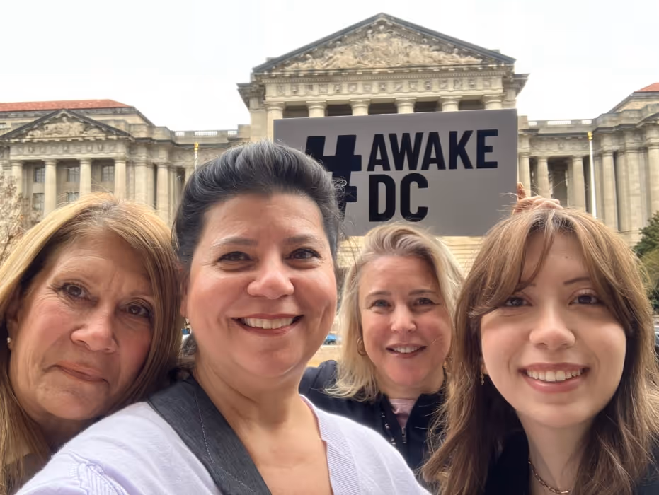 Four women take a cheerful group selfie outside a historic D.C. building, holding a large “#AWAKE DC” sign to highlight their participation in the event.