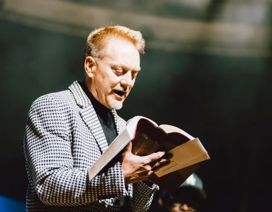 A speaker onstage reads passionately from an open Bible, illuminated by focused lighting while wearing a patterned jacket, creating a strong and reverent moment.