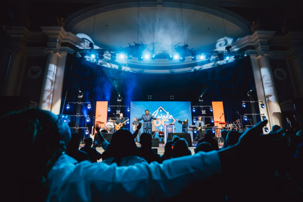 Worship on a brightly lit stage with the “Awake DC” backdrop, while attendees in the foreground raise their hands in praise under dramatic blue and white lighting.