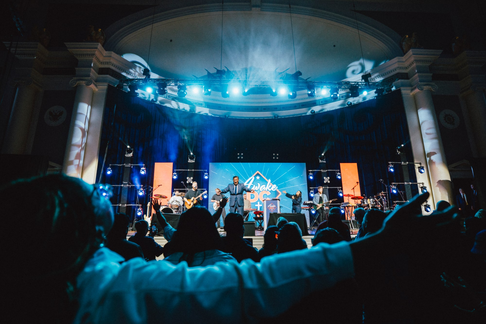 Worship on a brightly lit stage with the “Awake DC” backdrop, while attendees in the foreground raise their hands in praise under dramatic blue and white lighting.