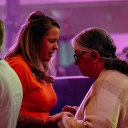 Two women standing close together, one holding the other’s hands, engaged in prayer or emotional support.