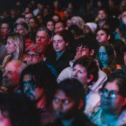 A large crowd of people seated closely together, listening attentively during a gathering, illuminated by mixed blue and red stage lighting.