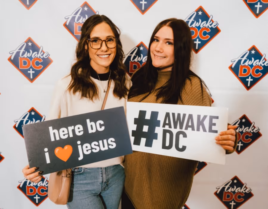 Two young women stand in front of an “Awake DC” event backdrop, smiling and holding signs. One sign reads “here bc I ❤️ jesus,” and the other displays “#AWAKE DC.” They appear engaged and enthusiastic, capturing the upbeat, faith-driven energy of the event