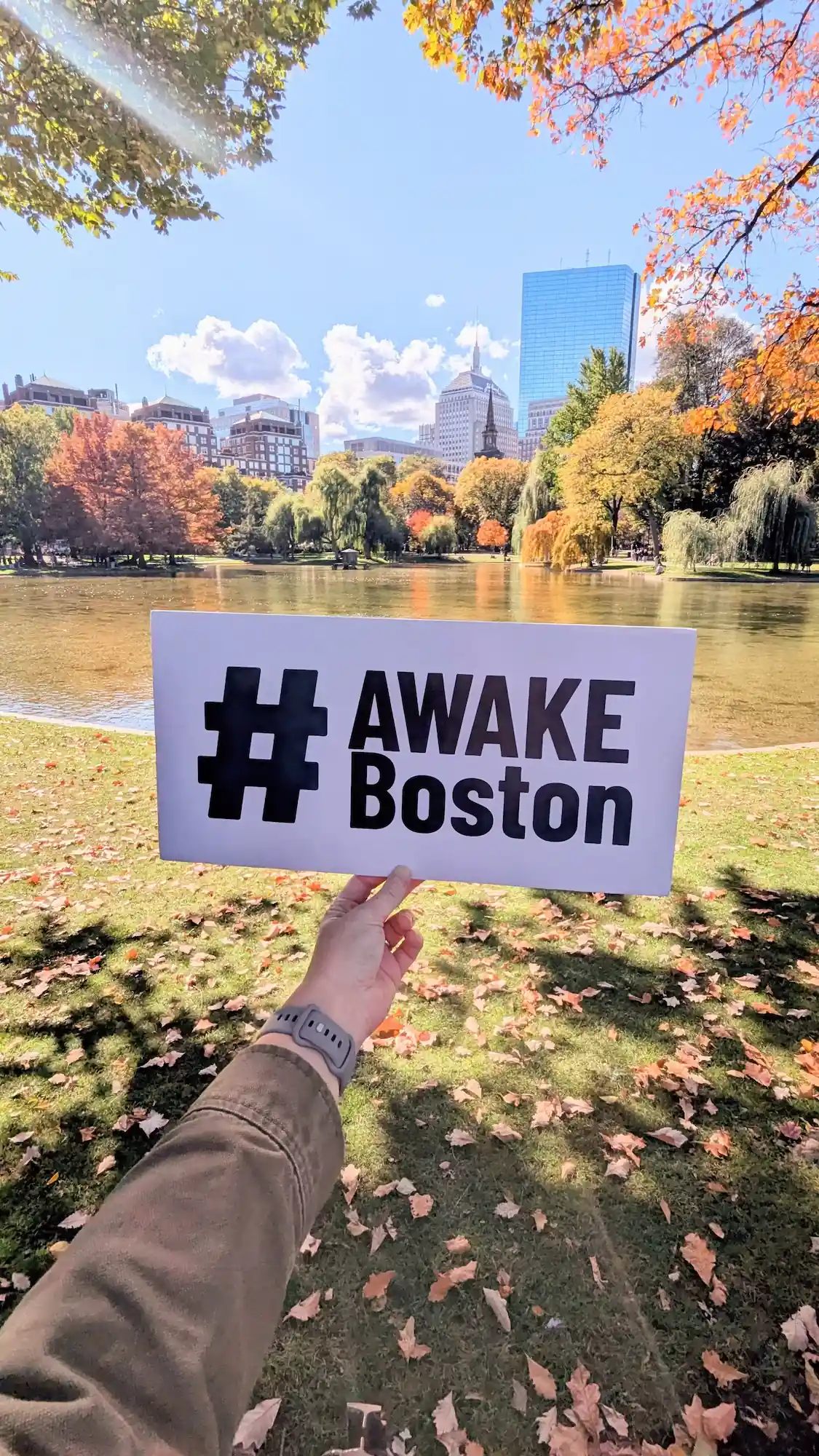 Hand holding #AWAKE Boston sign at Boston Public Garden with autumn fall foliage