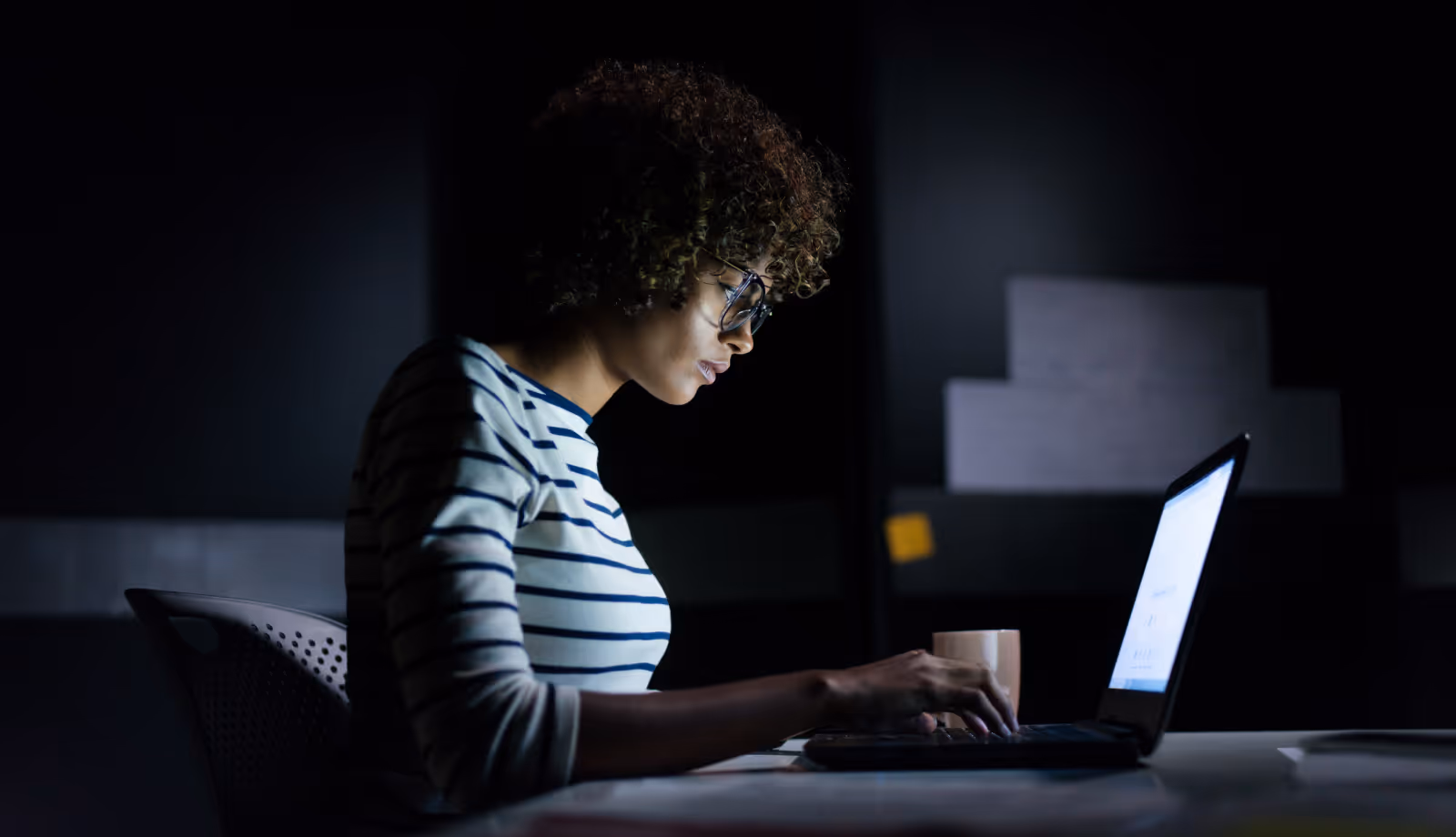 Person with curly hair and glasses working on a laptop in a dark room with a coffee cup nearby.