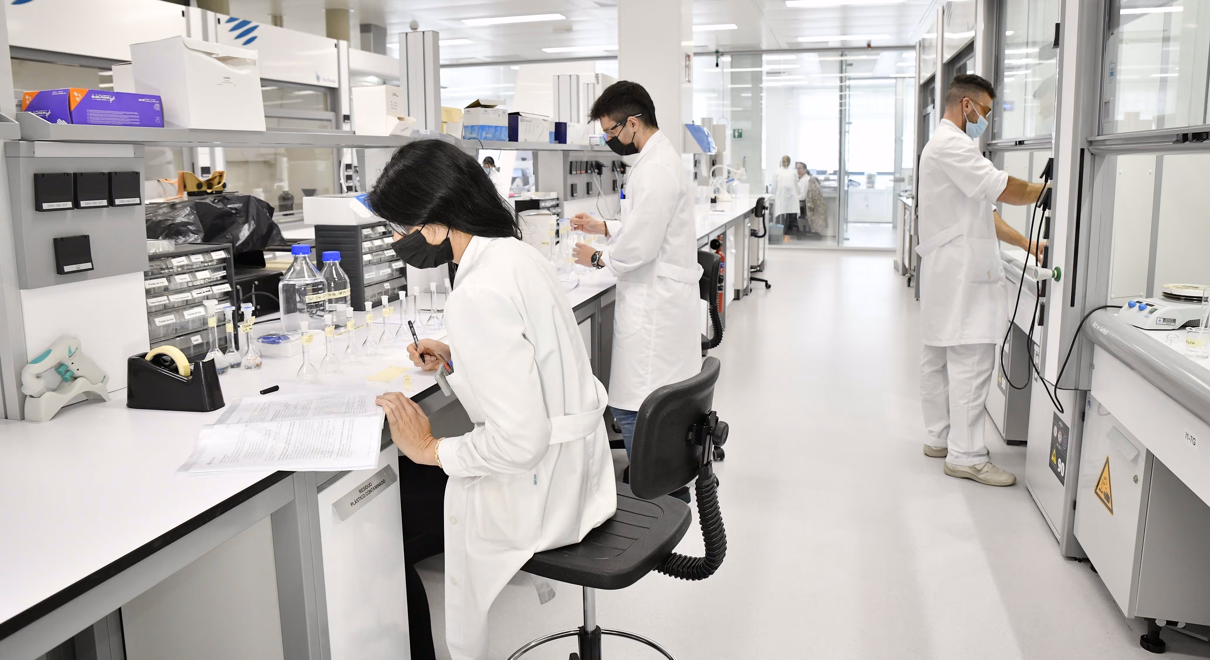 Researchers wearing white lab coats and face masks conducting experiments and recording notes in a modern laboratory.