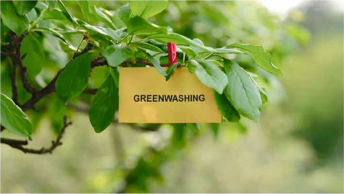 Close-up of green tree leaves with a yellow paper card clipped to a branch using a red wooden clothes peg. The card has the word "GREENWASHING" printed on it in bold, black capital letters against a blurred green background.