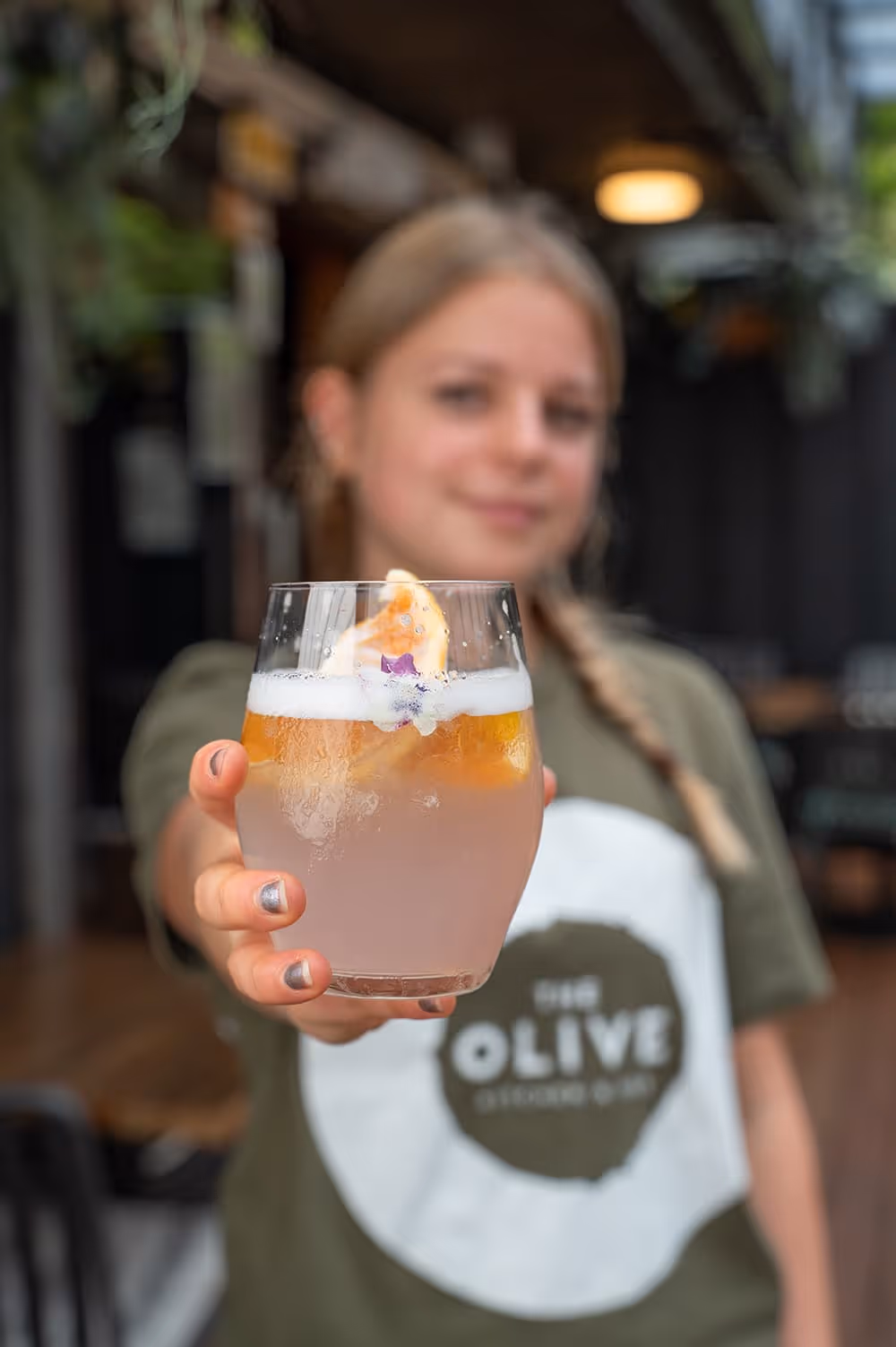 A team member at The Olive Kitchen and Bar holding a Lychee Sakura cocktail garnished with a fresh citrus slice and edible flowers