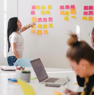 Person pointing at colorful sticky notes on a wall during a team meeting, with colleagues working at a table