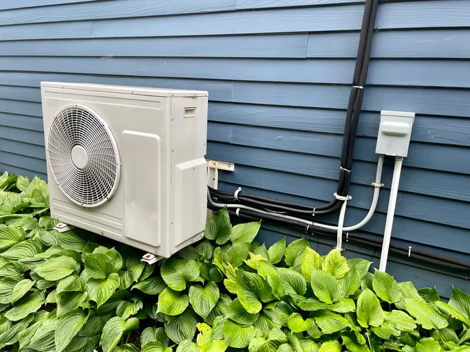 White air conditioning unit mounted on blue siding near green leafy plants