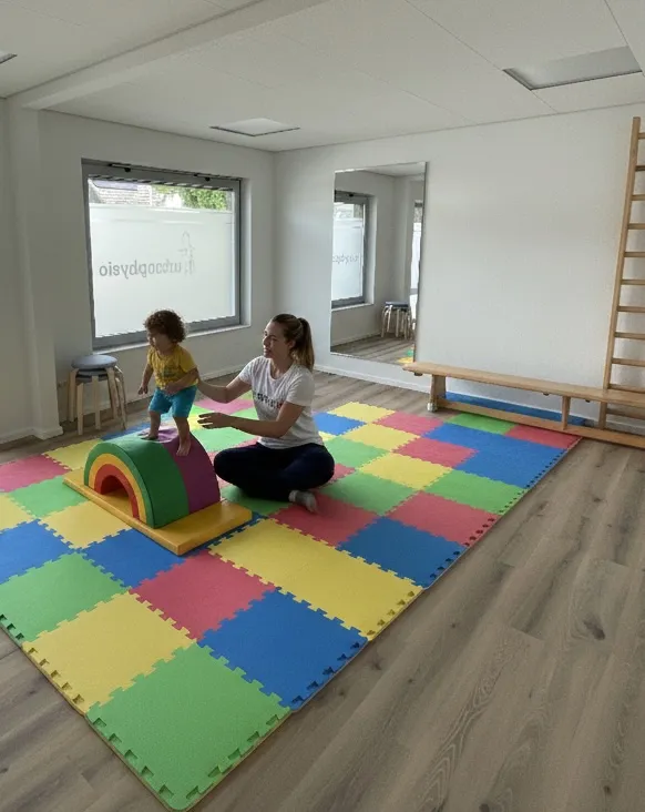 Ein Kind steht auf einem bunten Schaumstoff-Regenbogen-Schaukelstuhl, der von einer erwachsenen Frau auf einer mehrfarbigen Schaumstoffmatte in einem hellen Raum mit Holzboden und einem großen Spiegel gestützt wird.