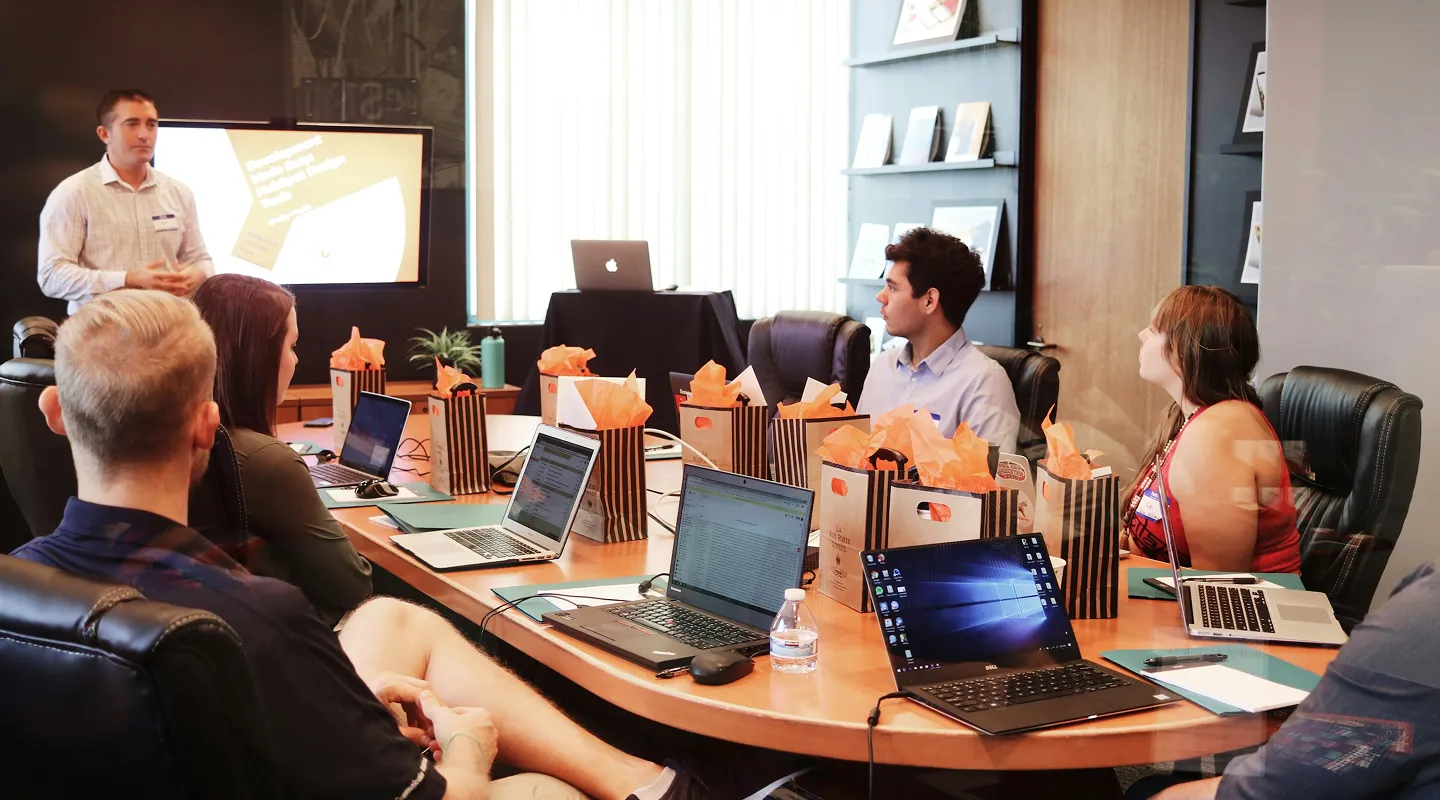 A photograph capturing a moment during a business presentation. A man is speaking to a group of colleagues gathered around a conference table, with a presentation screen in the background. The atmosphere suggests a professional and collaborative environment.