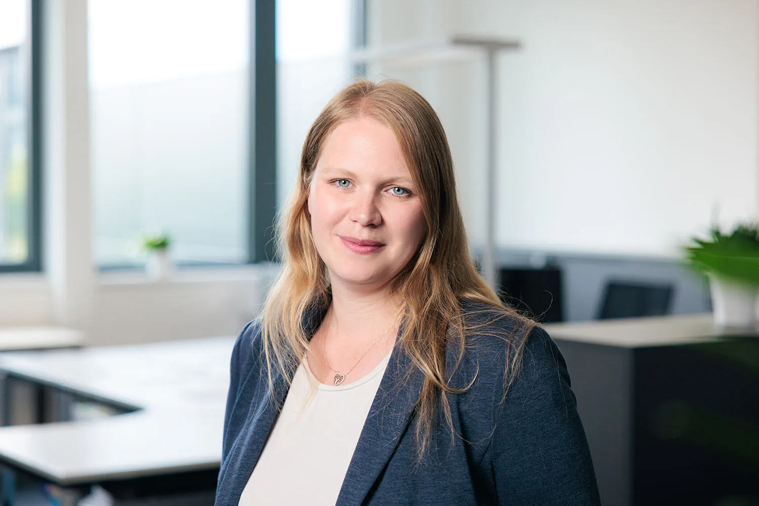 Porträt einer Frau mit schulterlangen blonden Haaren und dunklem Blazer vor Büroarbeitsplätzen.