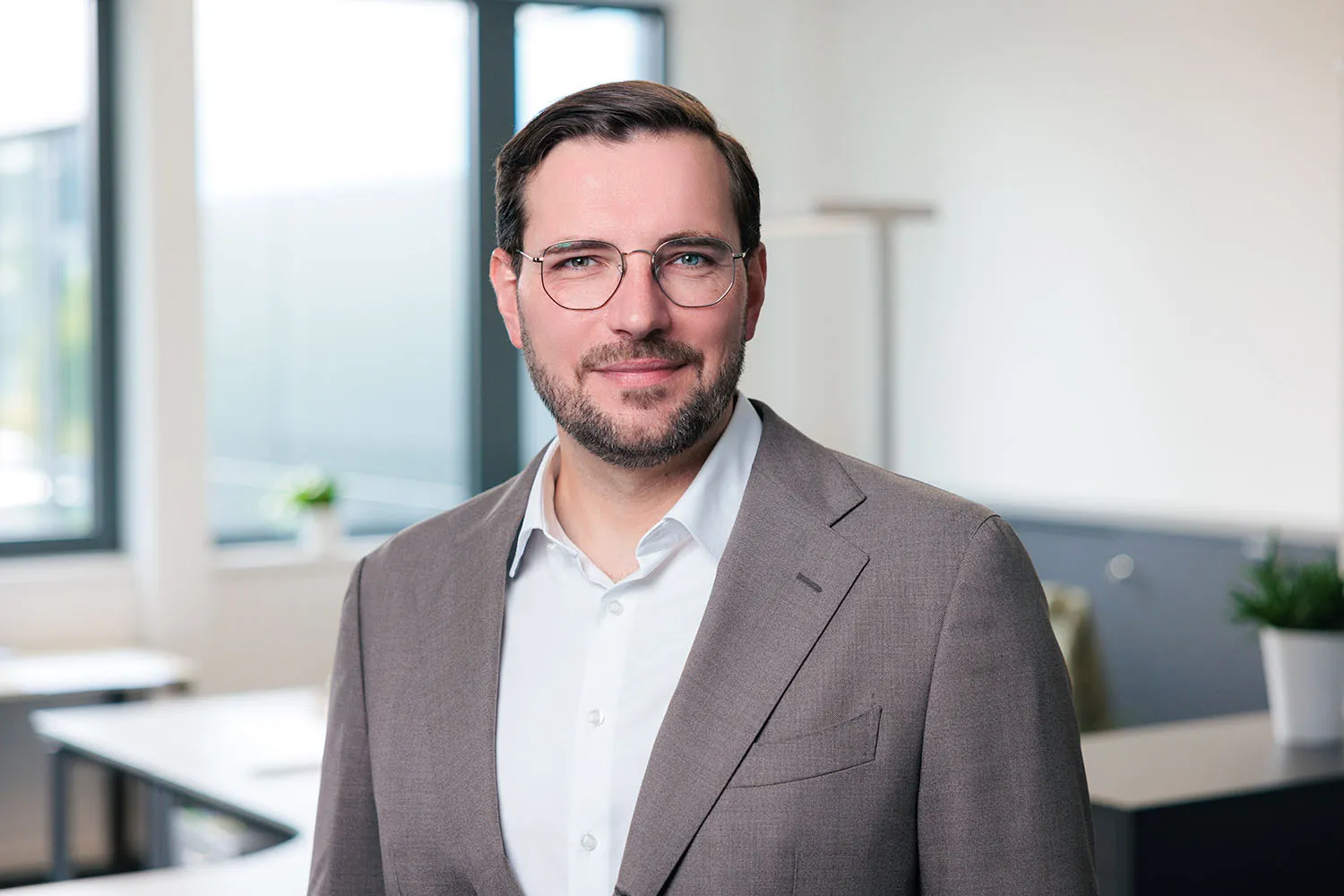 Porträt eines Mannes mit Bart, Brille und grauem Sakko vor modernem Büro.