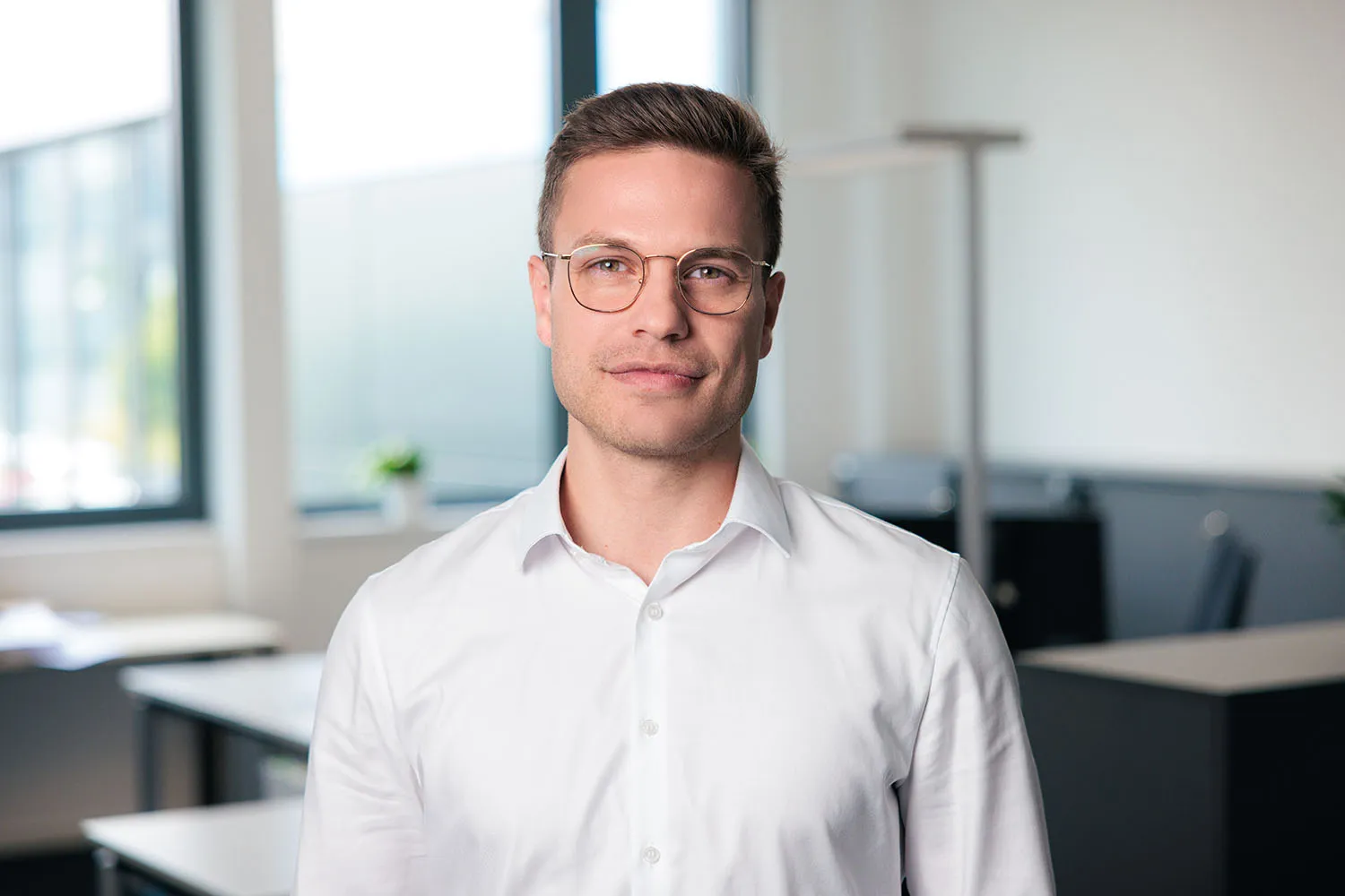 Porträt eines Mannes mit Brille, weißem Hemd und neutralem Gesichtsausdruck vor Büroarbeitsplätzen.