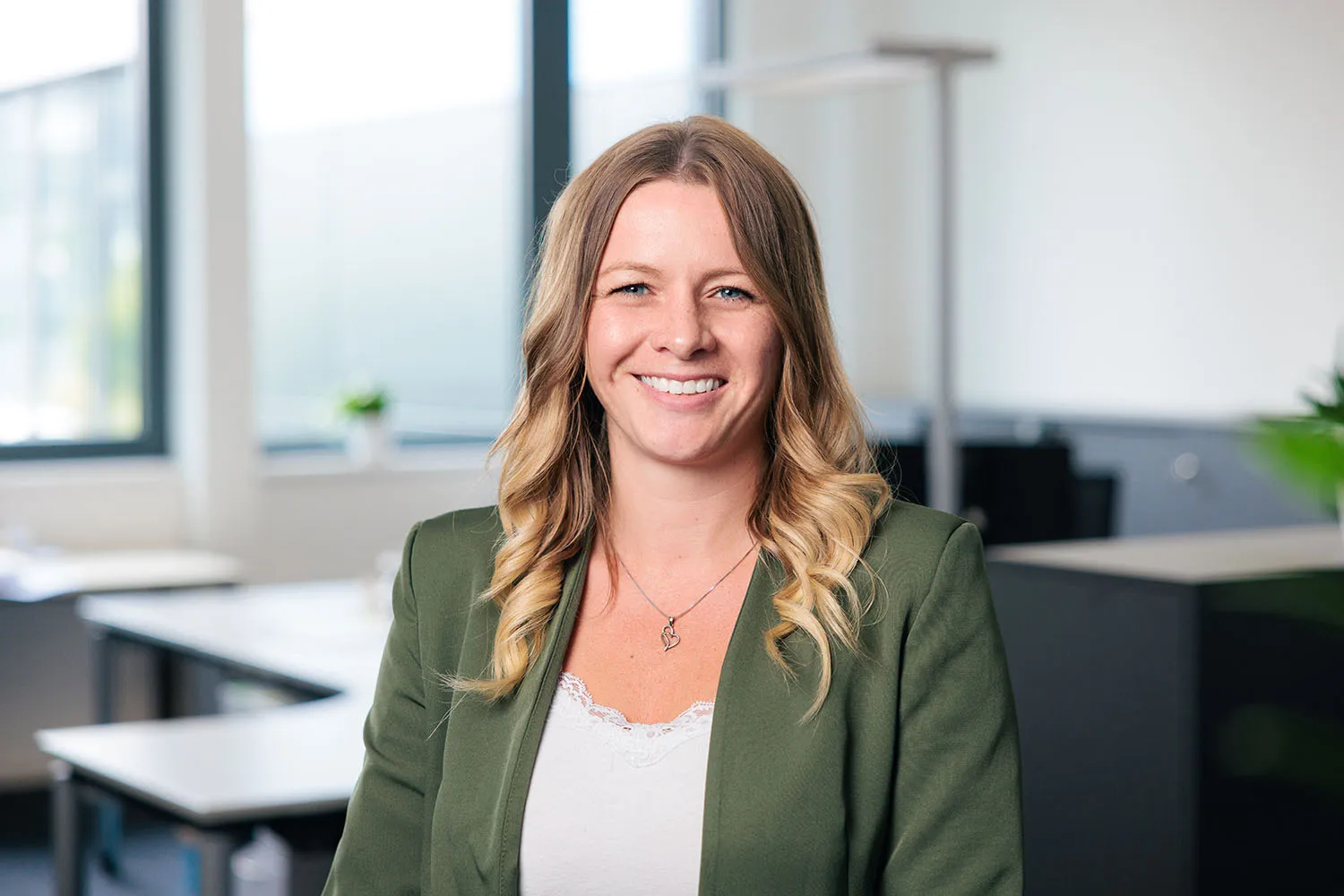 Porträt einer Frau mit blonden Haaren, grünem Blazer und hellem Oberteil, lächelnd im Büro.