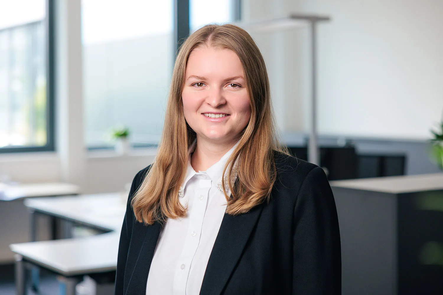 Porträt einer Frau mit schulterlangen Haaren, schwarzem Blazer und hellem Oberteil im Büro.