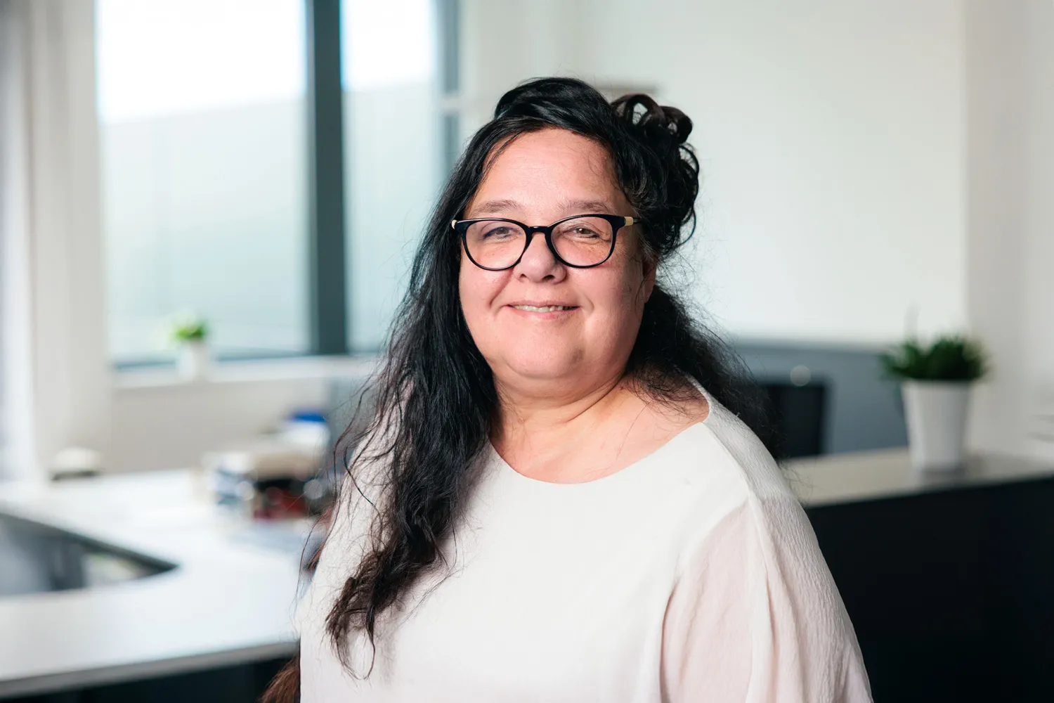 Porträt einer Frau mit dunklen Haaren und Brille, helles Oberteil, vor Bürohintergrund.