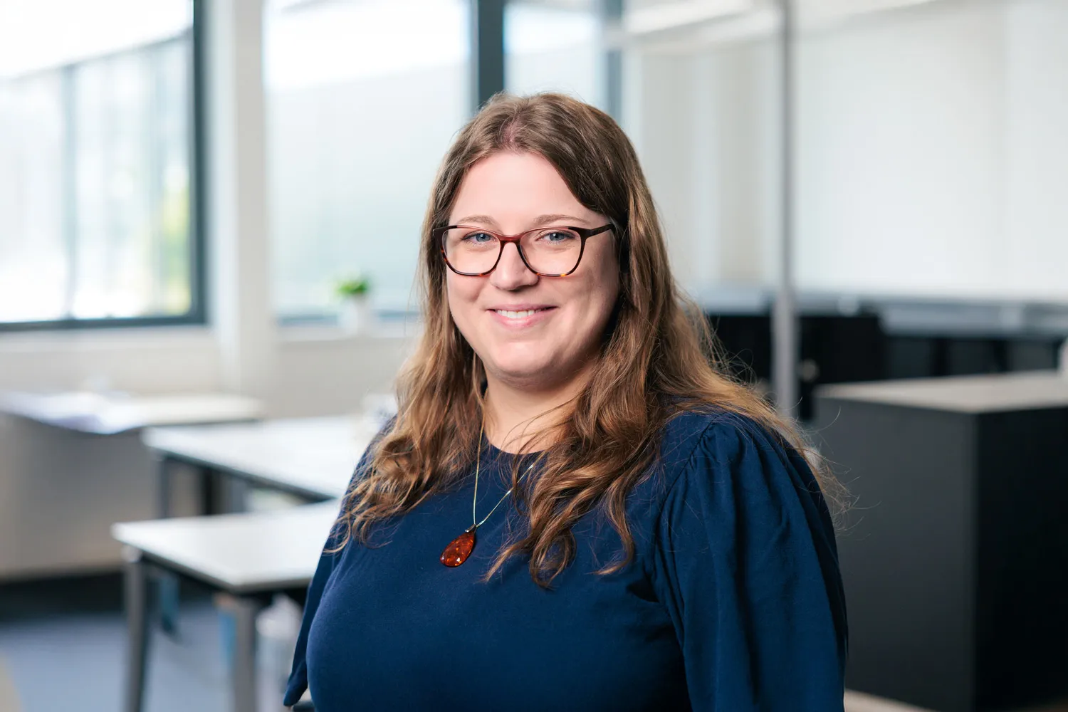 Porträt einer Frau mit Brille und dunklem Oberteil, lächelnd vor Bürohintergrund.