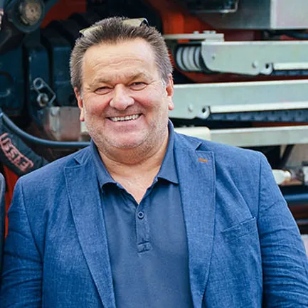 Mann in blauer Jacke lächelnd in einer industriellen Umgebung.