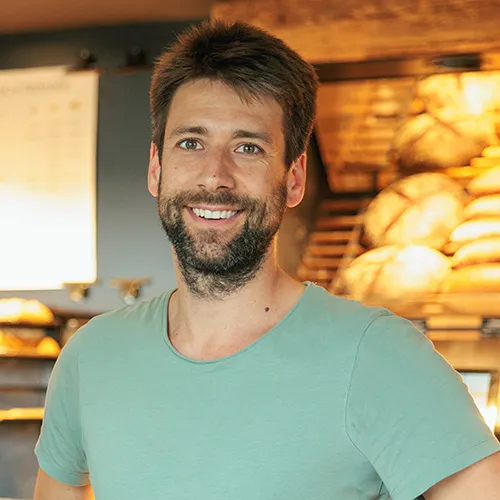 Mann mit Bart und grünem T-Shirt, lächelnd vor einem Regal mit Backwaren.