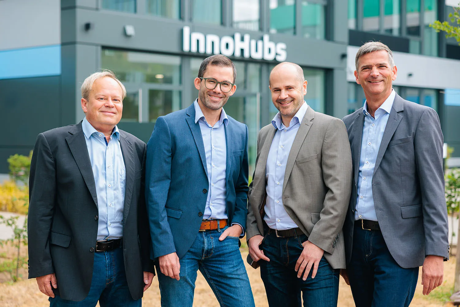 Gruppe von fünf Männern im Anzug, stehend vor einem modernen Bürogebäude im Außenbereich.