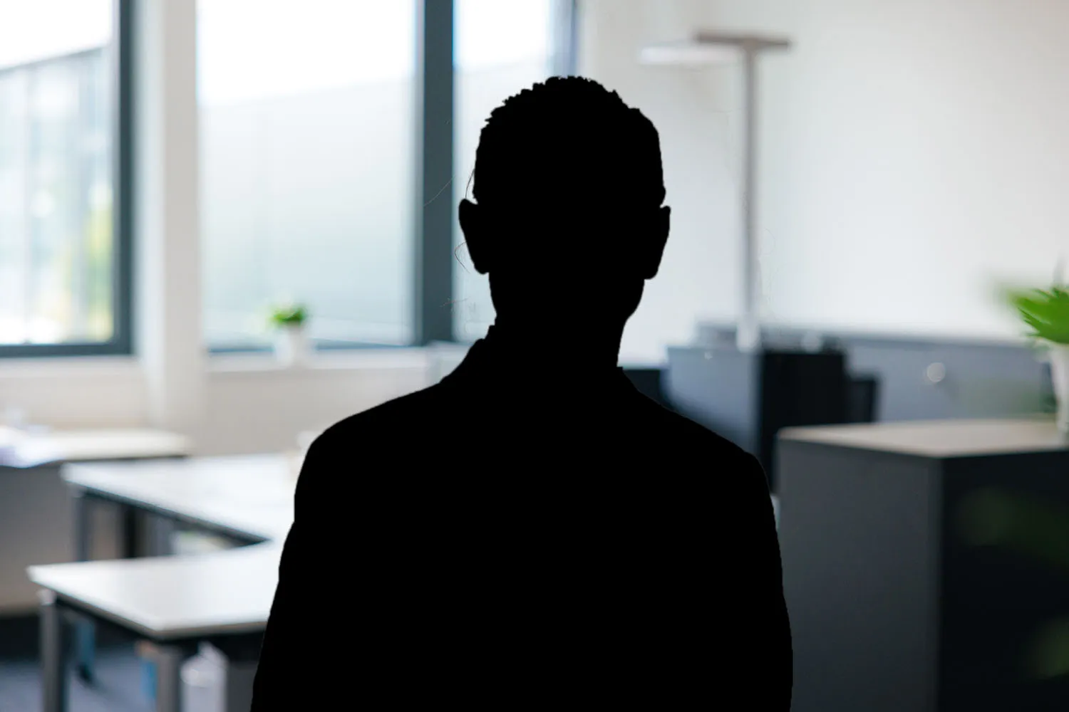 Silhouette einer sitzenden Person vor einem Schreibtisch in hellem Büro, Person nicht erkennbar.