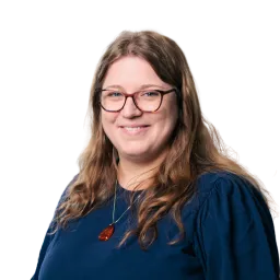 Porträt einer Frau mit Brille, langen Haaren und blauem Oberteil vor dunklem Hintergrund.