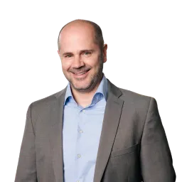 Porträt eines Mannes mit Glatze, grauem Sakko und Hemd, lächelnd vor dunklem Hintergrund.