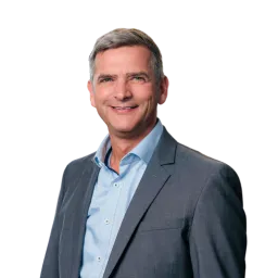 Porträt eines Mannes mit grauen Haaren, blauem Sakko und Hemd vor dunklem Hintergrund.