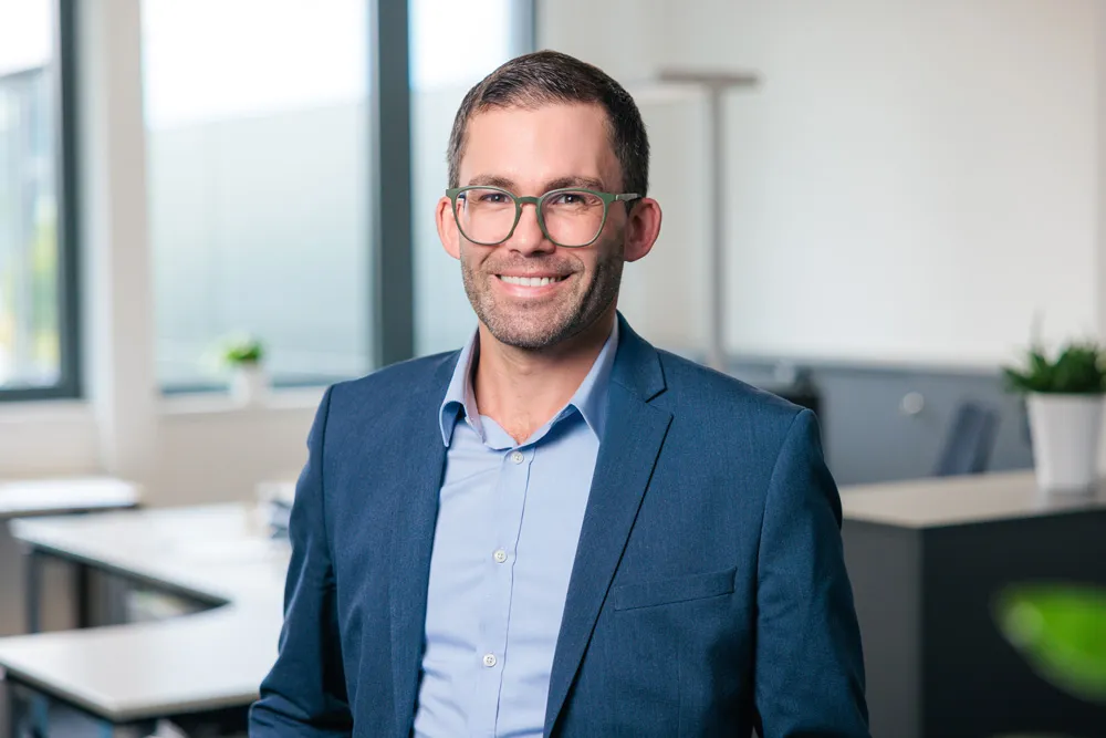 Porträt eines Mannes mit Brille, Bart und blauem Sakko, stehend vor einem hellen Büro.