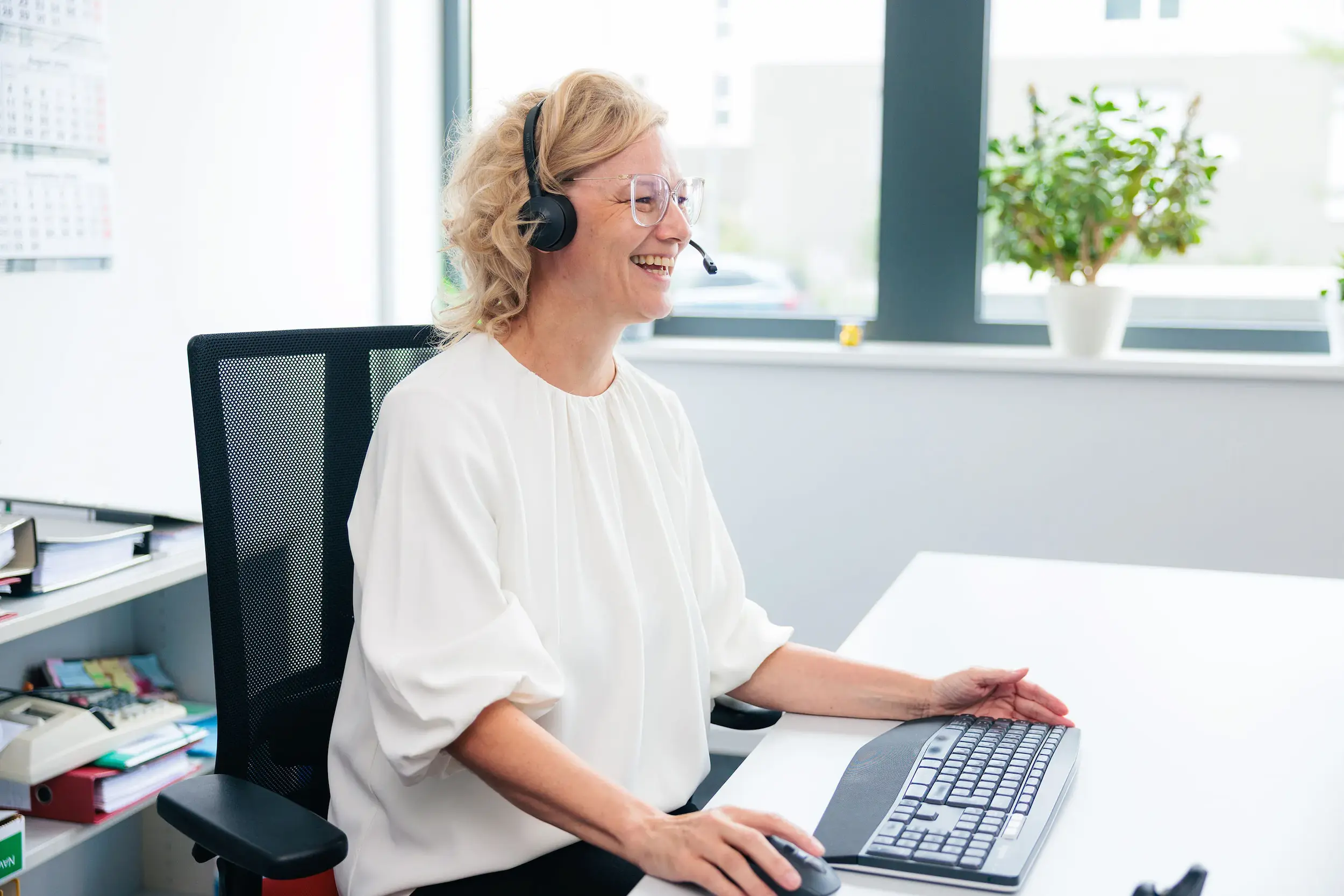 Lächelnde Frau im Büro mit Headset sitzt an einem Schreibtisch und arbeitet mit Tastatur und Maus.