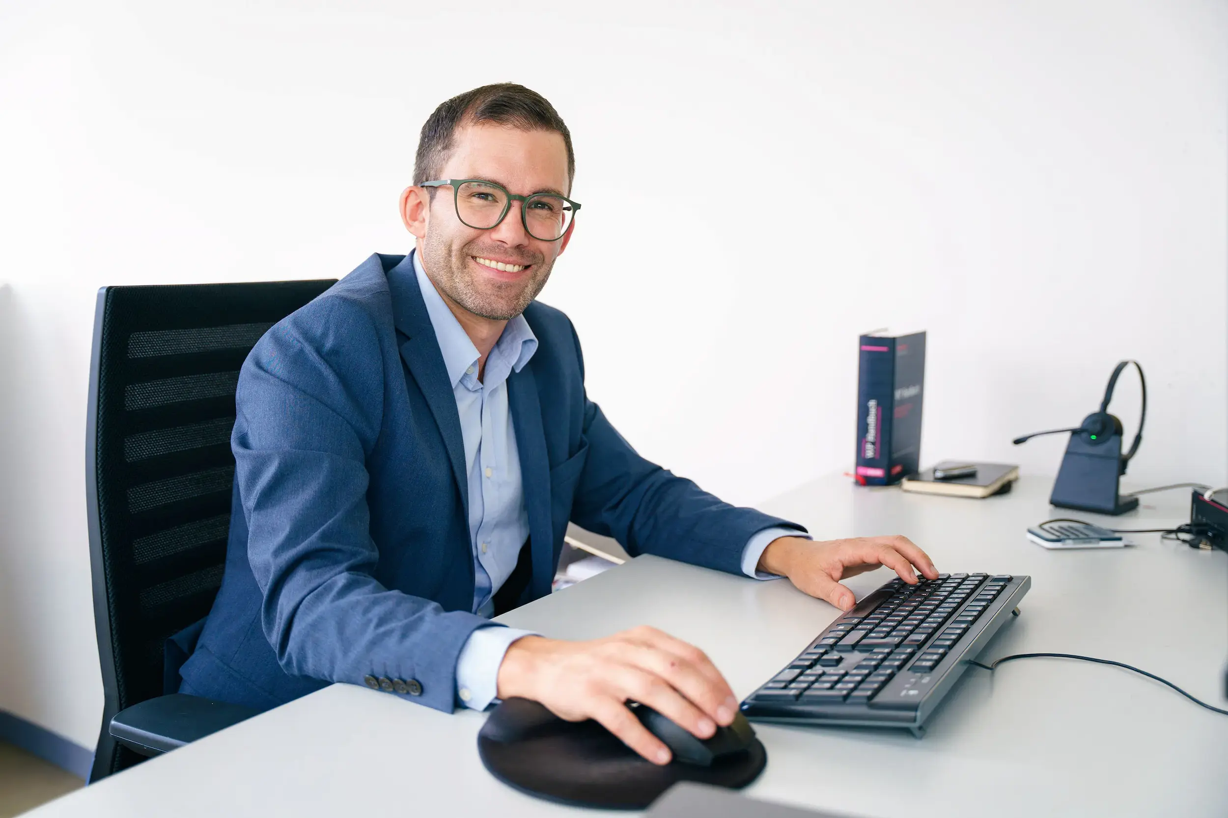 Lächelnder Mann in Brille und blauem Anzug sitzt am Schreibtisch mit Tastatur und Maus.