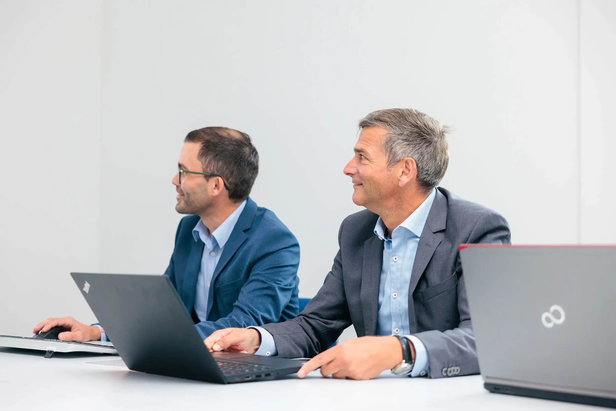 Zwei Männer im Anzug sitzen an einem weißen Tisch mit geöffneten Laptops und blicken nach links, einem Meeting folgend.