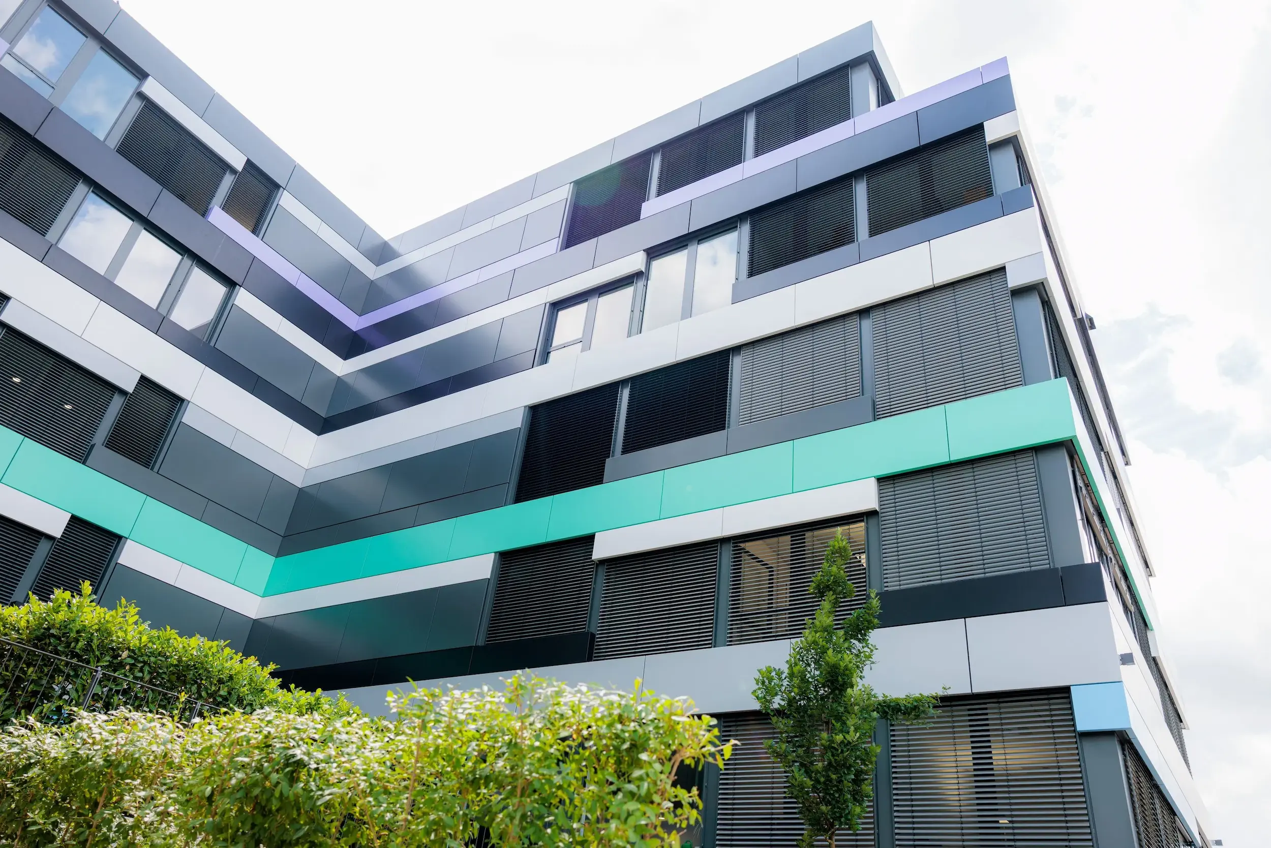 Moderne Bürogebäudeecke mit grauer Fassadenverkleidung, grünen Akzentstreifen und Fensterjalousien.