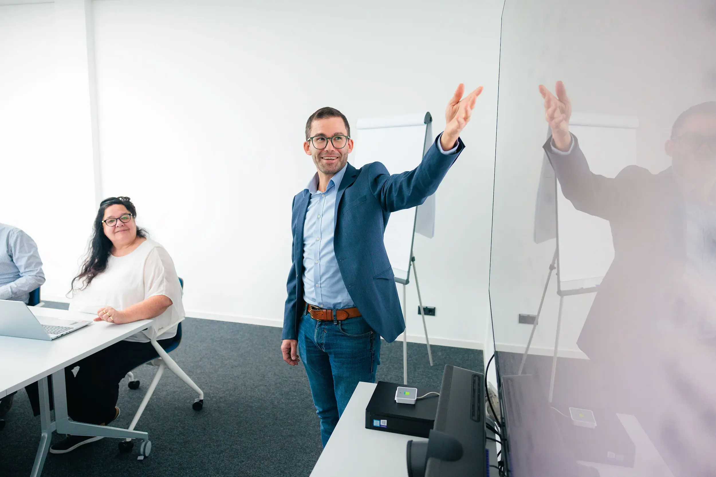 Ein Mann im blauen Sakko präsentiert in einem Konferenzraum vor einer Gruppe, während eine Frau an einem Tisch mit Laptop sitzt und ihm zuhört.