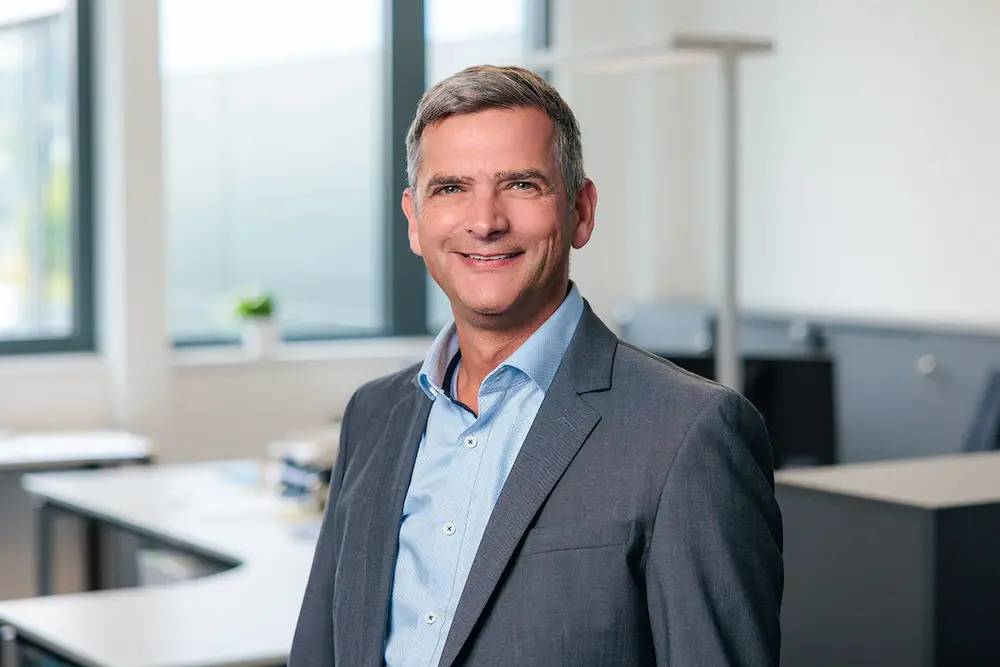 Mann mittleren Alters in grauem Anzug und hellblauem Hemd lächelt in einem modernen Büro.