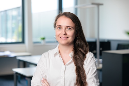Lächelnde Frau mit langen braunen Haaren und weißem Hemd vor einem modernen Bürofenster.